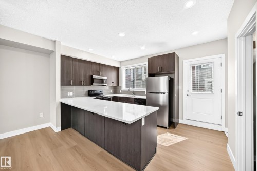 Kitchen featuring dark wood-finish cabinetry, stainless steel appliances, light countertops, a tile backsplash, and light wood-finish flooring - #75 2803 James Mowatt Tr Sw, Edmonton, AB 