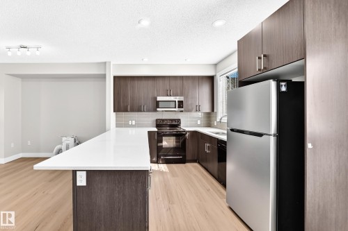 Modern kitchen featuring dark wood-finish cabinetry, stainless steel appliances, a white countertop island, and wood-finish flooring - #75 2803 James Mowatt Tr Sw, Edmonton, AB 