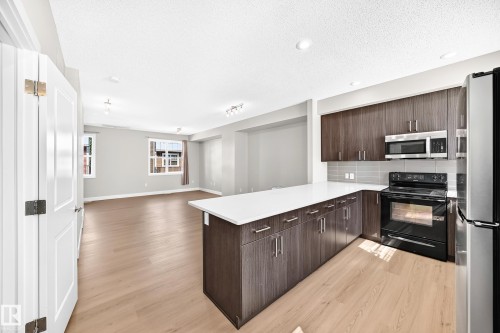 Open-concept kitchen and living area featuring wood-finish flooring, white countertops, dark wood-finish cabinetry, and stainless steel appliances - #75 2803 James Mowatt Tr Sw, Edmonton, AB 