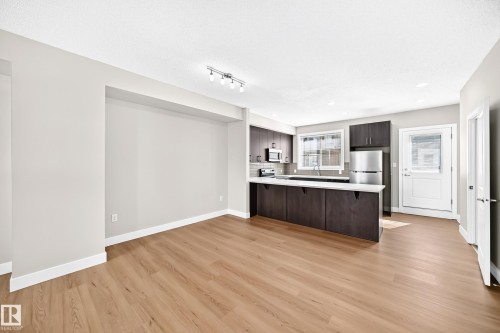 Open-concept living space with wood-finish flooring, a kitchen island, track lighting, and a full suite of stainless steel appliances - #75 2803 James Mowatt Tr Sw, Edmonton, AB 