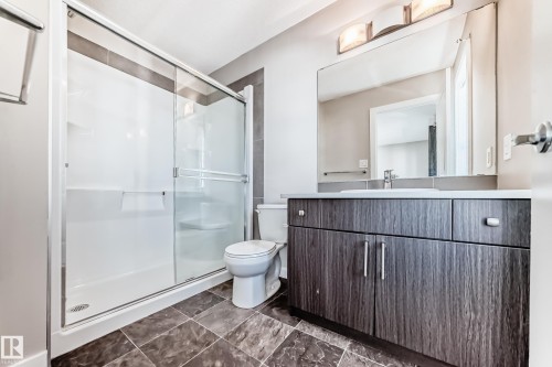 Framed shower with sliding glass door, single vanity with integrated sink, wood-finish cabinetry, and a large wall-mounted mirror - #75 2803 James Mowatt Tr Sw, Edmonton, AB 