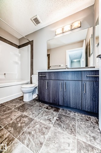 Bathroom featuring a full-length vanity mirror, a three-light fixture, wood-finish cabinetry, a white bathtub with a tiled surround, and textured floor tiling - #75 2803 James Mowatt Tr Sw, Edmonton, AB 