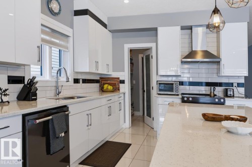 Modern kitchen featuring white glossy cabinetry, light-toned countertops, a stainless steel range hood, subway tile backsplash, and a glass-paneled pantry door - 527 Merlin Landing Nw, Edmonton, AB - Indoor Photo Showing Kitchen