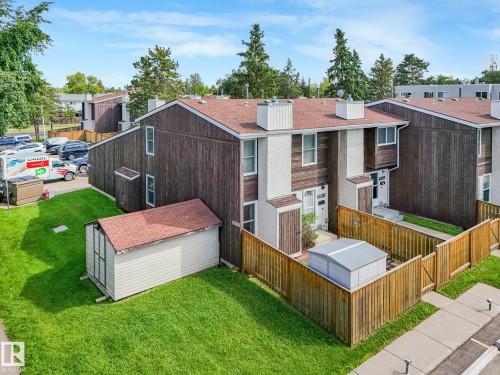 Wood-paneled townhouse exterior with a pitched shingle roof, private fenced yard, grass lawn, and a separate storage shed - 18417 66 Avenue Nw, Edmonton, AB - Outdoor With Exterior