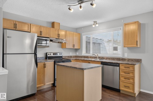 Kitchen featuring wood-finish cabinetry, stainless steel appliances, a center island with a stone-look countertop, and a double basin sink beneath a window - 6008 South Terwillegar Boulevard Nw, Edmonton, AB - Indoor Photo Showing Kitchen