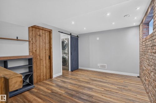 Wood-finish flooring, exposed brick wall, recessed ceiling lighting, and a barn door leading to a laundry area with stacked appliances - 6008 South Terwillegar Boulevard Nw, Edmonton, AB - Indoor Photo Showing Other Room