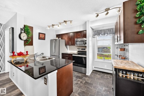Kitchen featuring dark wood-finish cabinetry, stainless steel appliances, black countertops, a dual basin sink, and slate-style floor tiling - #401 5390 Chappelle Rd Sw, Edmonton, AB 
