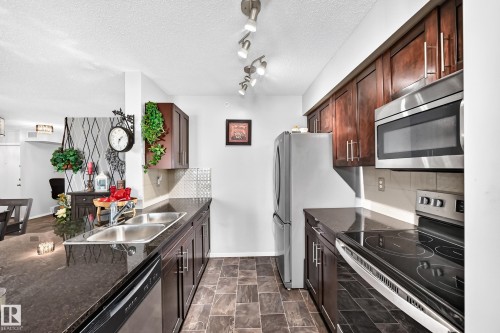 Kitchen featuring dark wood-finish cabinetry, stainless steel appliances, dark stone countertops, and tile flooring - #401 5390 Chappelle Rd Sw, Edmonton, AB 