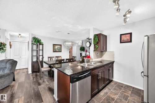 Kitchen island with dark countertops, stainless steel sink, and built-in dishwasher - #401 5390 Chappelle Rd Sw, Edmonton, AB 