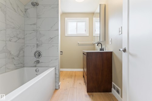 White bathtub with subway tile surround, wood-finish flooring, dark wood-tone vanity, white countertop, and rectangular window - 12337 128 Avenue Nw, Edmonton, AB - Indoor Photo Showing Bathroom