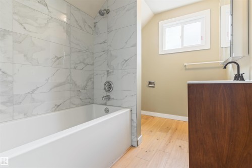 Bathtub with marble-look tile surround, wood-finish vanity with countertop, and light wood-finish flooring - 12337 128 Avenue Nw, Edmonton, AB - Indoor Photo Showing Bathroom