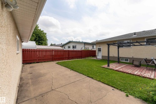 Expansive concrete driveway, manicured lawn, red picket fence, wood deck with pergola, and stucco exterior - 7120 139 Avenue Nw, Edmonton, AB - Outdoor