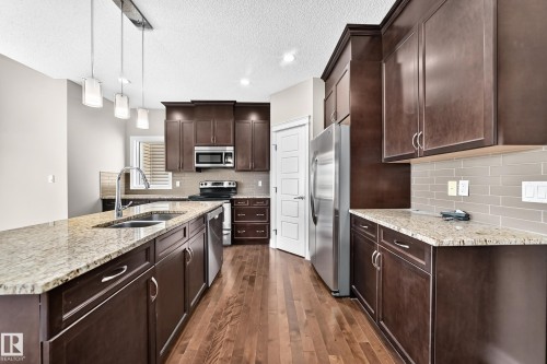 Kitchen featuring a large island with an undermount sink, pendant lighting, stainless steel appliances, dark wood-finish cabinetry, and light-toned countertops - 4212 Charles Close Sw, Edmonton, AB - Indoor Photo Showing Kitchen With Stainless Steel Kitchen With Double Sink With Upgraded Kitchen