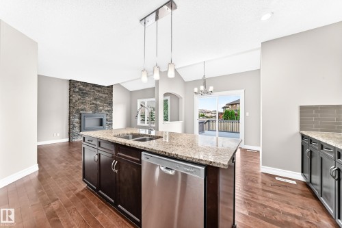Spacious kitchen island with granite countertop and stainless steel appliances, wood-finish flooring, stone-clad fireplace, vaulted ceiling, and sliding glass doors to outdoor deck space - 4212 Charles Close Sw, Edmonton, AB - Indoor Photo Showing Kitchen With Double Sink With Upgraded Kitchen