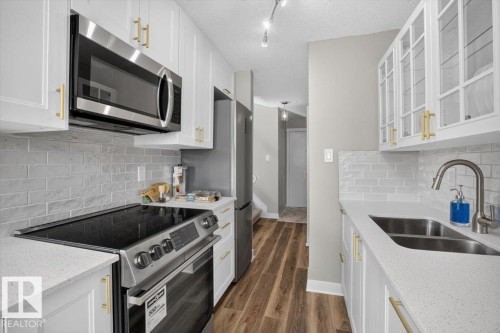 White cabinetry with gold-tone hardware, stainless steel appliances, light-toned countertops, subway tile backsplash, and wood-finish flooring - #205 11429 124 St Nw, Edmonton, AB - Indoor Photo Showing Kitchen With Double Sink With Upgraded Kitchen