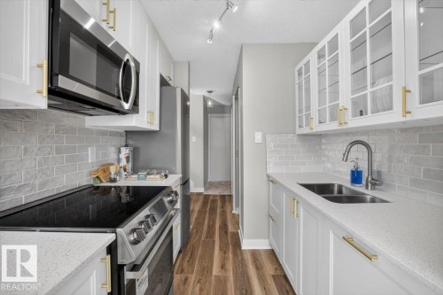 White cabinetry with gold-tone hardware, stainless steel appliances, light gray subway tile backsplash, light-toned countertops, and wood-finish flooring - #205 11429 124 St Nw, Edmonton, AB - Indoor Photo Showing Kitchen With Double Sink With Upgraded Kitchen