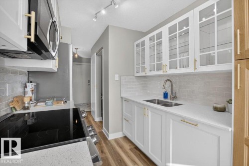 Kitchen featuring white shaker cabinetry, glass-front upper cabinets, light wood-finish flooring, a stainless steel double basin sink, and white subway tile backsplash - #205 11429 124 St Nw, Edmonton, AB - Indoor Photo Showing Kitchen With Double Sink With Upgraded Kitchen