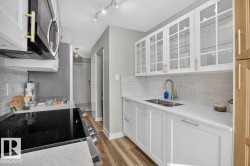 Kitchen featuring white shaker cabinetry, glass-front upper cabinets, light wood-finish flooring, a stainless steel double basin sink, and white subway tile backsplash - 