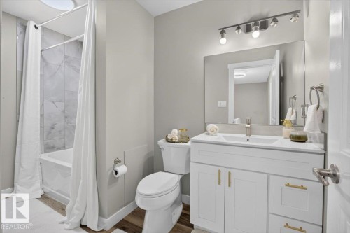 Bathroom featuring a built-in bathtub with tiled surround, wood-finish flooring, a white vanity with gold-tone hardware, and a rectangular mirror with track lighting - #205 11429 124 St Nw, Edmonton, AB - Indoor Photo Showing Bathroom
