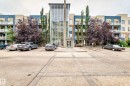 Multi-story building featuring a prominent glass-enclosed central entrance, multiple balconies with metal railings, and a parking area - #102 2584 Anderson Way Sw, Edmonton, AB  - Outdoor With Facade 