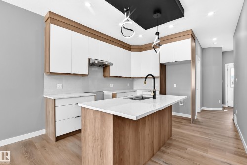 Kitchen featuring wood-finish flooring, white cabinetry with wood accents, a white countertop island with an integrated sink, a contemporary black faucet, and pendant lighting fixtures - 426 Crystal Creek Link, Leduc, AB - Indoor Photo Showing Kitchen With Double Sink