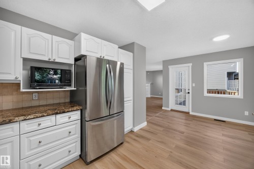 White cabinetry with brushed nickel hardware, stone-look countertops with a tile backsplash, stainless steel refrigerator, light wood-finish flooring, and a glass-paneled door - 3640 31 Street Nw, Edmonton, AB - Indoor Photo Showing Kitchen With Stainless Steel Kitchen
