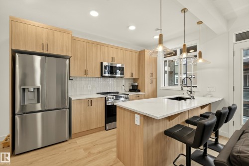 Modern kitchen featuring wood-finish cabinetry, stainless steel appliances, a white tile backsplash, and a large center island with a black sink and white countertop - #53 16231 19 Av Sw, Edmonton, AB - Indoor Photo Showing Kitchen With Upgraded Kitchen