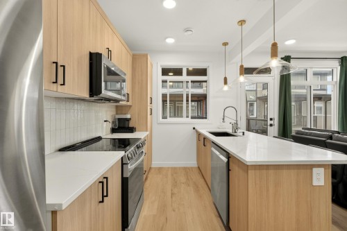 Contemporary kitchen featuring light wood-finish cabinetry with black hardware, stainless steel appliances, a white tile backsplash, and light-toned countertops - #53 16231 19 Av Sw, Edmonton, AB - Indoor Photo Showing Kitchen With Upgraded Kitchen