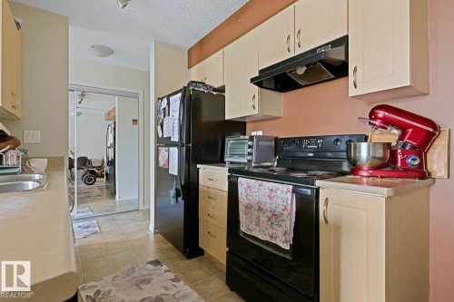 Galley-style kitchen featuring light-toned cabinetry, a black range with an overhead hood, a black refrigerator, and a stainless steel sink - #106 13907 136 St Nw, Edmonton, AB - Indoor Photo Showing Kitchen With Double Sink