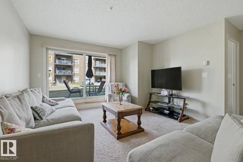 Living area featuring neutral wall tones, carpeted flooring, and a sliding glass door leading to an outdoor patio - #106 13907 136 St Nw, Edmonton, AB - Indoor Photo Showing Living Room