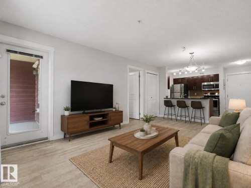 Open-concept living area with wood-finish flooring, a kitchen featuring dark wood cabinetry, stainless steel appliances, and a breakfast bar - #205 5816 Mullen Pl Nw, Edmonton, AB - Indoor Photo Showing Living Room