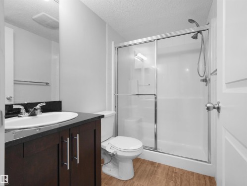 Bathroom featuring a dark wood-finish vanity with an undermount sink, a dark countertop, a walk-in shower with a sliding glass door, and wood-finish flooring - #205 5816 Mullen Pl Nw, Edmonton, AB - Indoor Photo Showing Bathroom