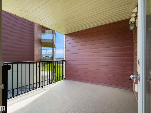 Covered balcony with a dark red siding wall, light-colored overhead soffit, and a black metal railing - #205 5816 Mullen Pl Nw, Edmonton, AB - Outdoor With Balcony With Exterior