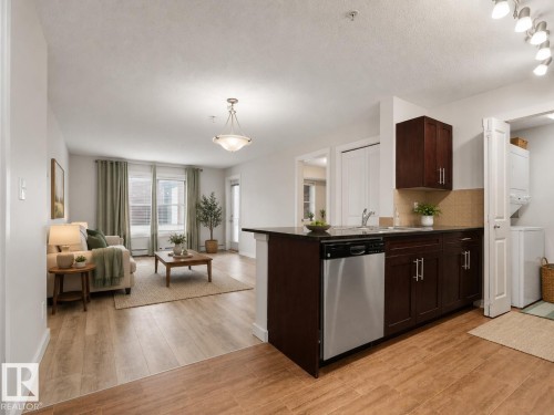Open-concept living area featuring wood-finish flooring, a kitchen island with dark cabinetry, stainless steel dishwasher, and track lighting - #205 5816 Mullen Pl Nw, Edmonton, AB - Indoor Photo Showing Kitchen