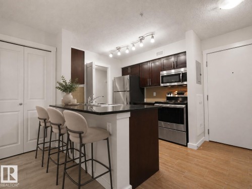Kitchen featuring a breakfast bar with dark countertops, stainless steel appliances, dark wood-finish cabinetry, and wood-finish flooring - #205 5816 Mullen Pl Nw, Edmonton, AB - Indoor Photo Showing Kitchen