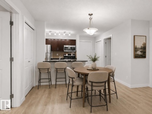 Open-concept layout featuring wood-finish flooring, a kitchen with dark cabinetry, stainless steel appliances, and a breakfast bar, complemented by a dining area with an overhead pendant light - #205 5816 Mullen Pl Nw, Edmonton, AB - Indoor Photo Showing Dining Room