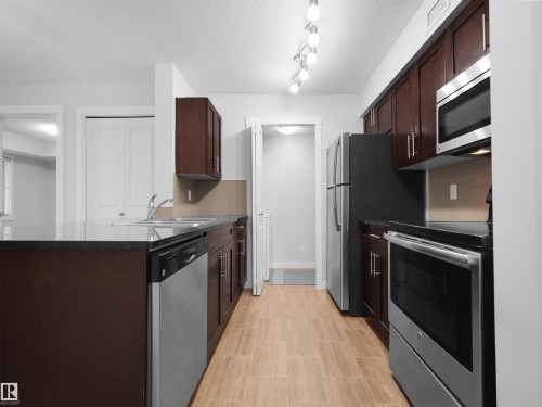 Kitchen featuring dark wood-finish cabinetry, black countertops, stainless steel appliances, and wood-finish flooring - #205 5816 Mullen Pl Nw, Edmonton, AB - Indoor Photo Showing Kitchen With Stainless Steel Kitchen