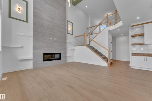 High ceilings with recessed lighting, linear fireplace with a stone-look surround, wood-finish flooring, glass railing staircase with wood accents, and built-in white cabinetry with wood shelving - 764 Astoria Way, Devon, AB - Indoor With Fireplace