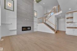 High ceilings with recessed lighting, linear fireplace with a stone-look surround, wood-finish flooring, glass railing staircase with wood accents, and built-in white cabinetry with wood shelving - 