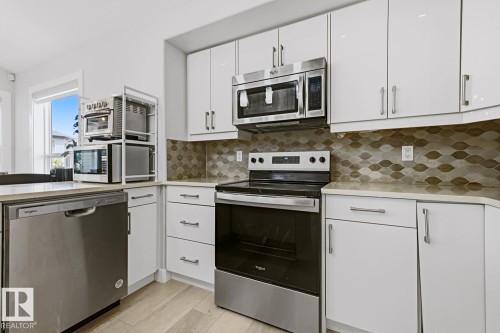 White upper and lower cabinetry, stainless steel appliances, patterned backsplash, light-toned countertops, and wood-finish flooring - 11816 168 Avenue Nw, Edmonton, AB - Indoor Photo Showing Kitchen With Upgraded Kitchen