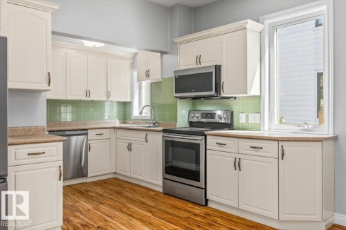 Kitchen featuring white cabinetry, light green tiled backsplash, stainless steel appliances, wood-finish flooring, and a large window - 11429 64 Street Nw, Edmonton, AB - Indoor Photo Showing Kitchen With Stainless Steel Kitchen