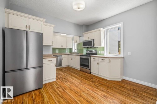 Kitchen featuring wood-finish flooring, light-toned cabinetry, stainless steel appliances, and a green tile backsplash - 11429 64 Street Nw, Edmonton, AB - Indoor Photo Showing Kitchen With Stainless Steel Kitchen