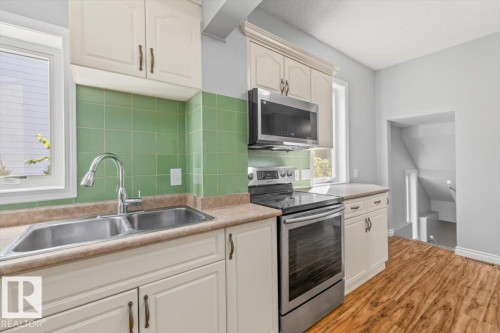 Kitchen featuring white cabinetry, stainless steel appliances, a double basin sink with a high-arc faucet, green tile backsplash, and wood-finish flooring - 11429 64 Street Nw, Edmonton, AB - Indoor Photo Showing Kitchen With Double Sink