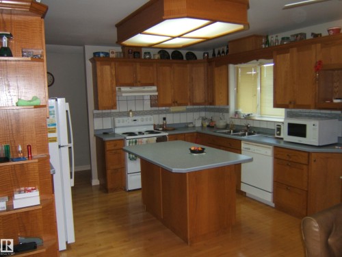 Kitchen featuring wood cabinetry, a central island with a solid surface countertop, white appliances, and wood-finish flooring - 5404 49 Street, Two Hills, AB - Indoor Photo Showing Kitchen With Double Sink