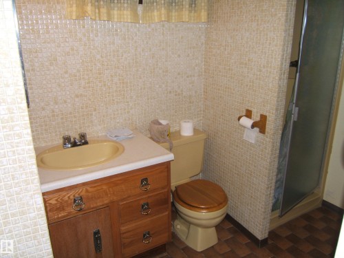 Wood-finish vanity with a built-in sink, tiled walls, and a glass-enclosed shower - 5404 49 Street, Two Hills, AB - Indoor Photo Showing Bathroom