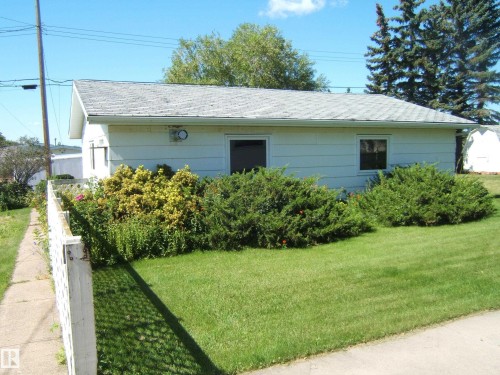 White siding exterior with a shingled roof, front yard with mature landscaping, and a white lattice fence - 5404 49 Street, Two Hills, AB - Outdoor