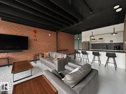 Open-concept living space with exposed brick walls, a modern kitchen island, dark cabinetry, stainless steel appliances, and sleek black pendant lighting - #301 9808 103 St Nw, Edmonton, AB - 