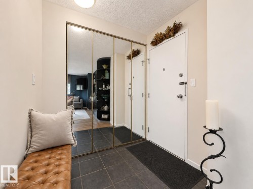Entryway featuring dark gray square tile flooring, a white entry door with chrome hardware, and full-height mirrored closet doors with brass trim - #301 9808 103 St Nw, Edmonton, AB - Indoor Photo Showing Other Room
