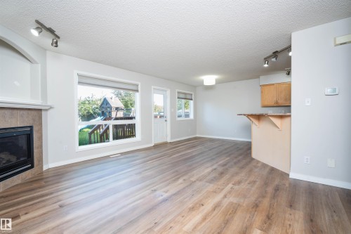 Spacious open concept room featuring wood-finish flooring, a tiled fireplace with mantel, multiple large windows, and track lighting - 1009 Macewan Close Sw, Edmonton, AB - Indoor Photo Showing Living Room With Fireplace