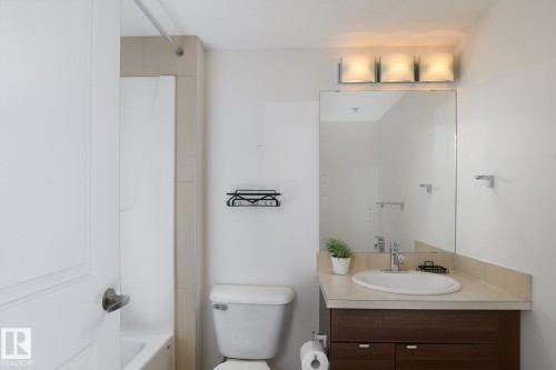 Bathroom featuring a wood-finish vanity with an integrated sink, a large wall-mounted mirror, and a three-light fixture - #110 9523 160 Av Nw, Edmonton, AB - Indoor Photo Showing Bathroom
